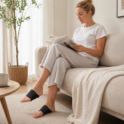 Kvinna sitter i en soffa och läser medan hon bär ergonomiska fotvalvsstöd med elastisk rem.